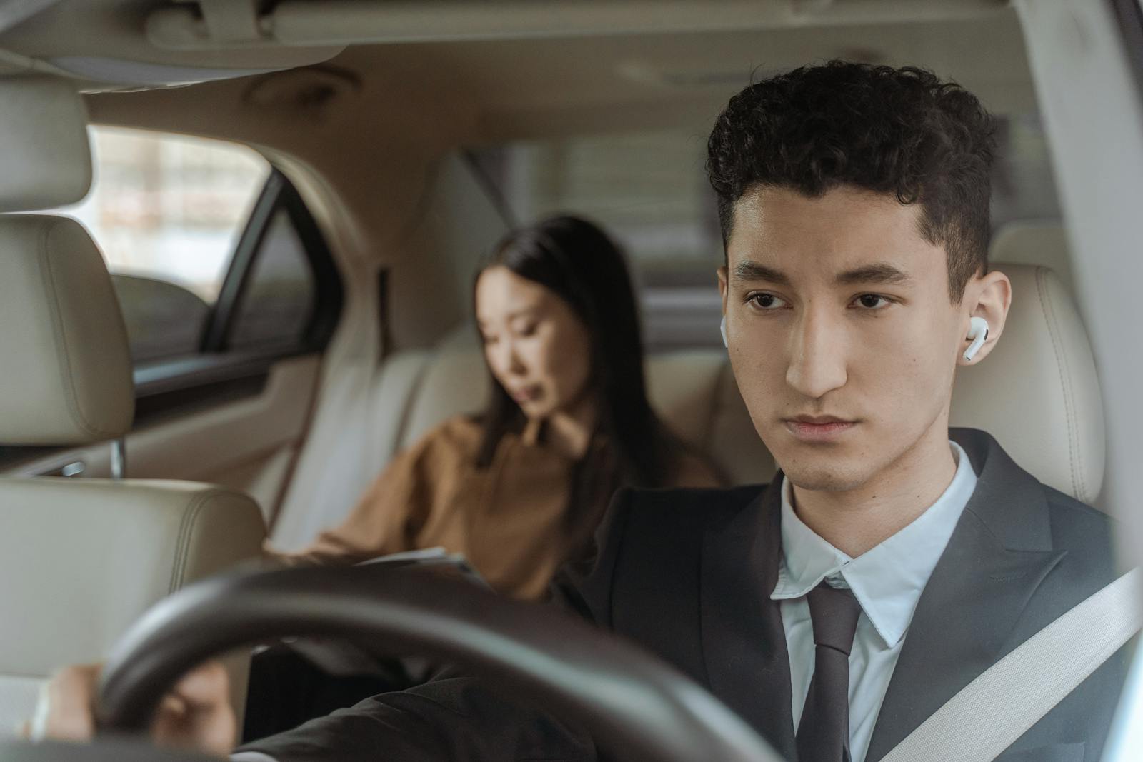 Young professional driver wearing suit and earpiece in luxury car interior with female passenger.